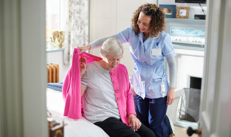 An older woman sat on a bed being helped by a carer to put a cardigan on