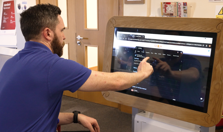 man with dark hair and a beard wearing a blue polo shirt sitting at a screen and pointing at information on it which from a distance looks like a care record