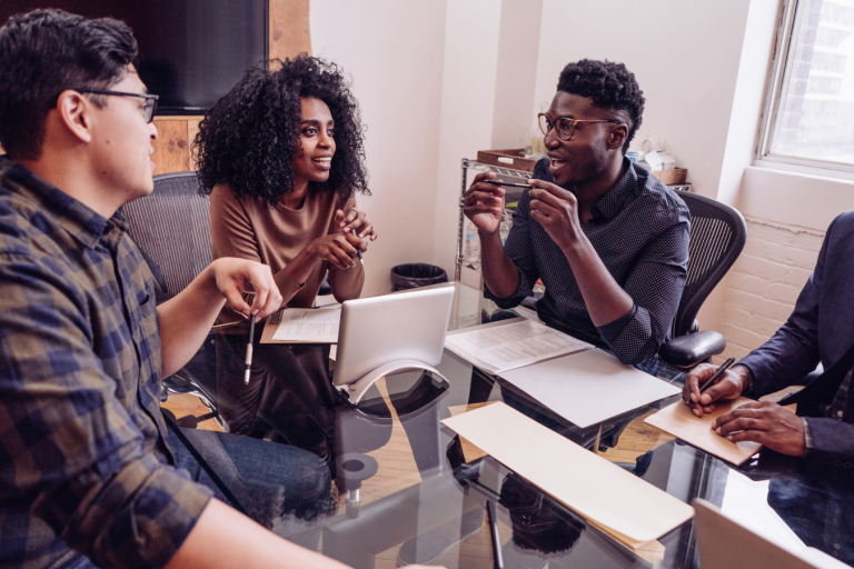 Four people sat around a table talking. There is a laptop and notebook on the table.