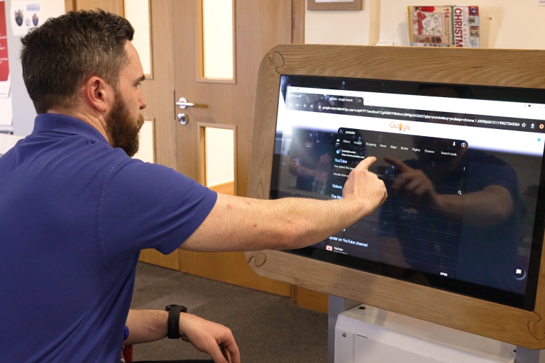 man with dark hair and a beard wearing a blue polo shirt sitting at a screen and pointing at information on it which from a distance looks like a care record